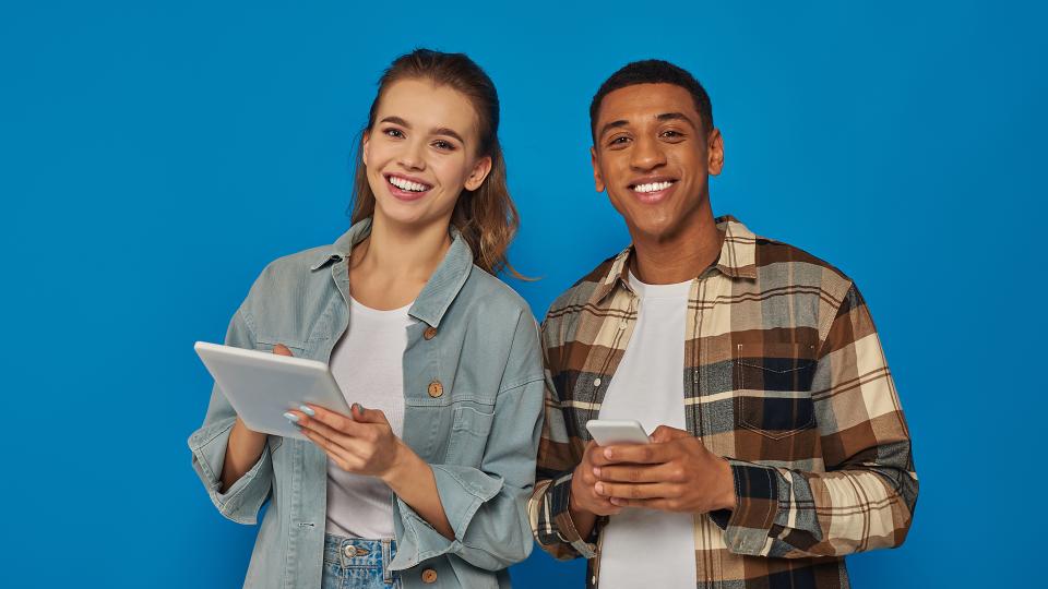 Von links: Junge Frau mit digitalem Tablet und junger Mann mit Smartphone vor blauem Hintergrund, beide lächeln in die Kamera