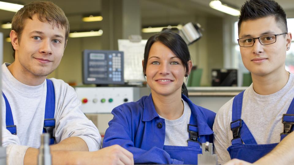 Drei Auszubildende in blauer Arbeitskleidung in Werkhalle