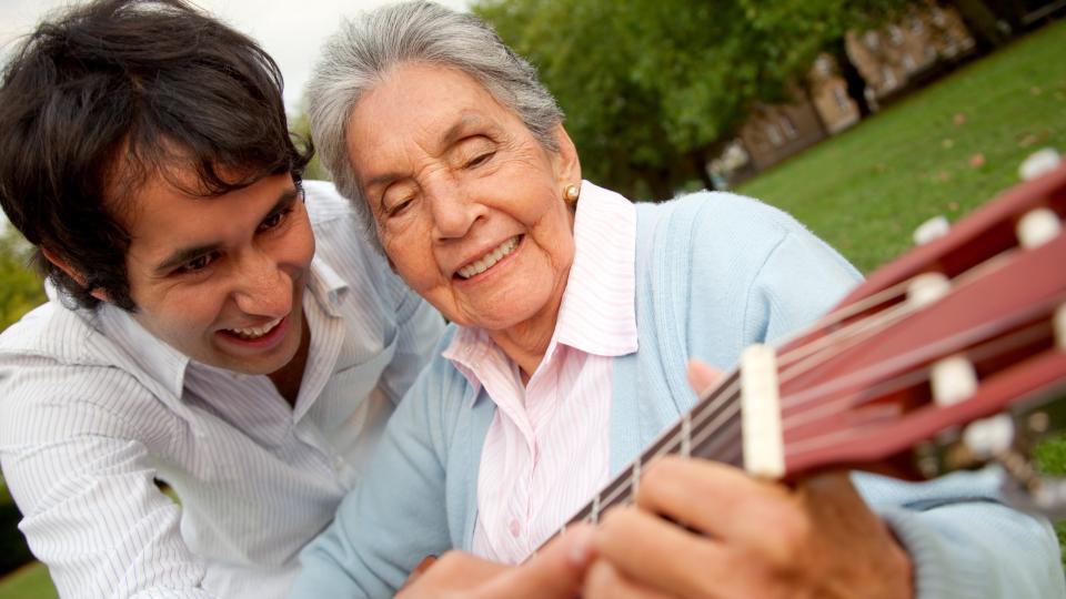 Ein jüngerer Gitarrenlehrer zeigt einer lebensalten Dame einen Gitarrengriff - beide lächeln.